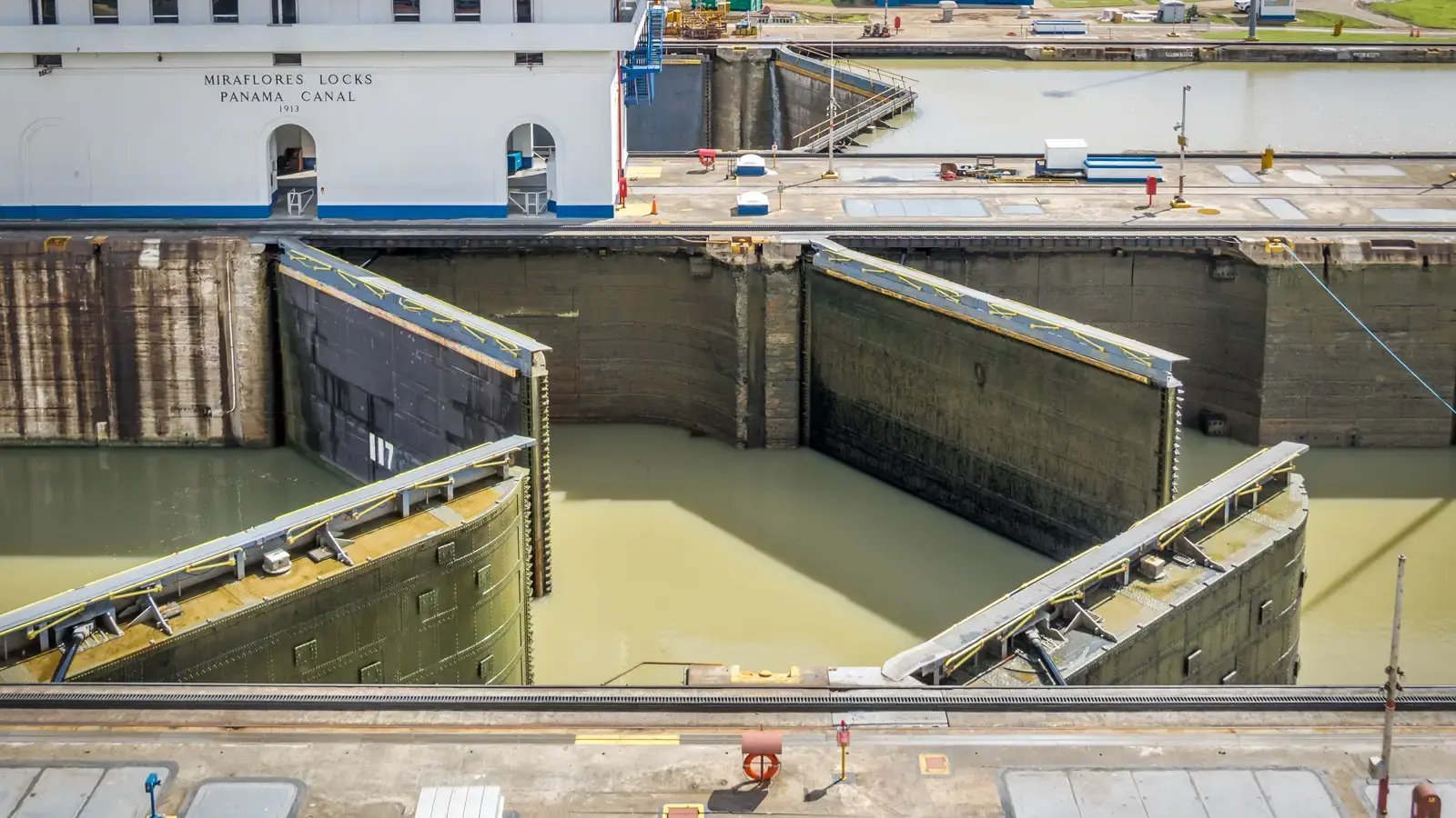 An elevated daytime view of the Miraflores Locks at the Panama Canal, showing the massive steel lock gates partially open as murky water flows between sections.