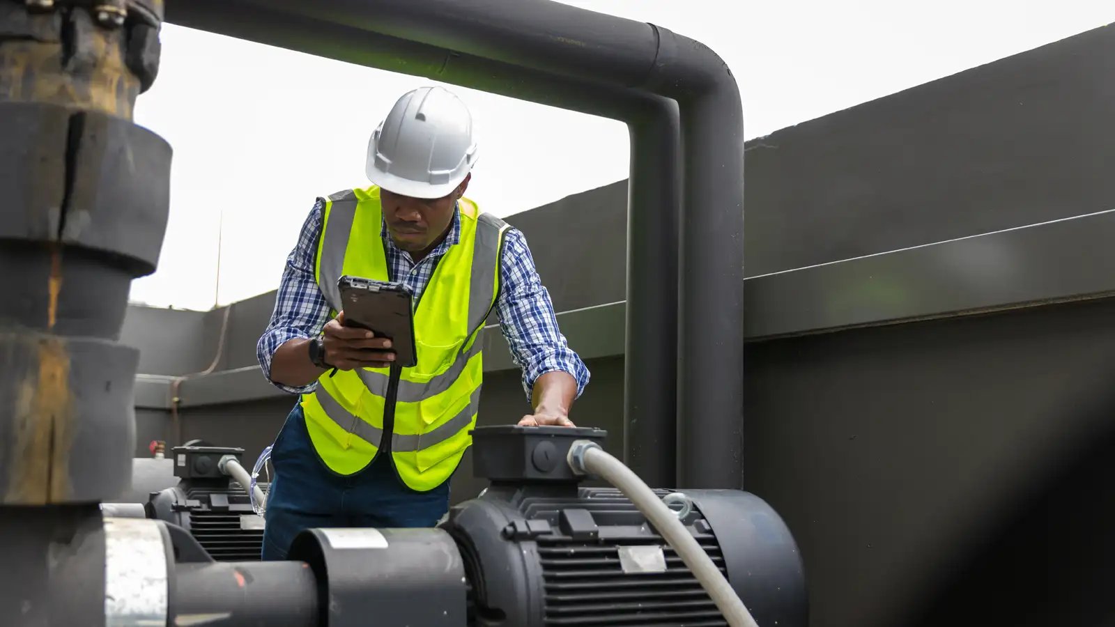 Technician in a hard hat and hi-vis vest inspecting industrial pumps on a rugged tablet during a smart monitoring check.